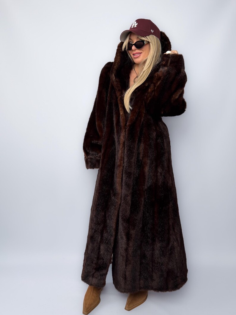 May include: A long, dark brown faux fur coat with a hood. The coat is floor-length and has a soft, textured appearance. The person is wearing a maroon baseball cap and sunglasses. The coat is a statement piece for cold weather.