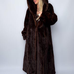 May include: A full-length, dark brown faux fur coat with a large hood. The coat has a long, flowing design and a plush texture. The hood is also lined with the same faux fur material. The coat is worn by a person wearing sunglasses and a baseball cap.