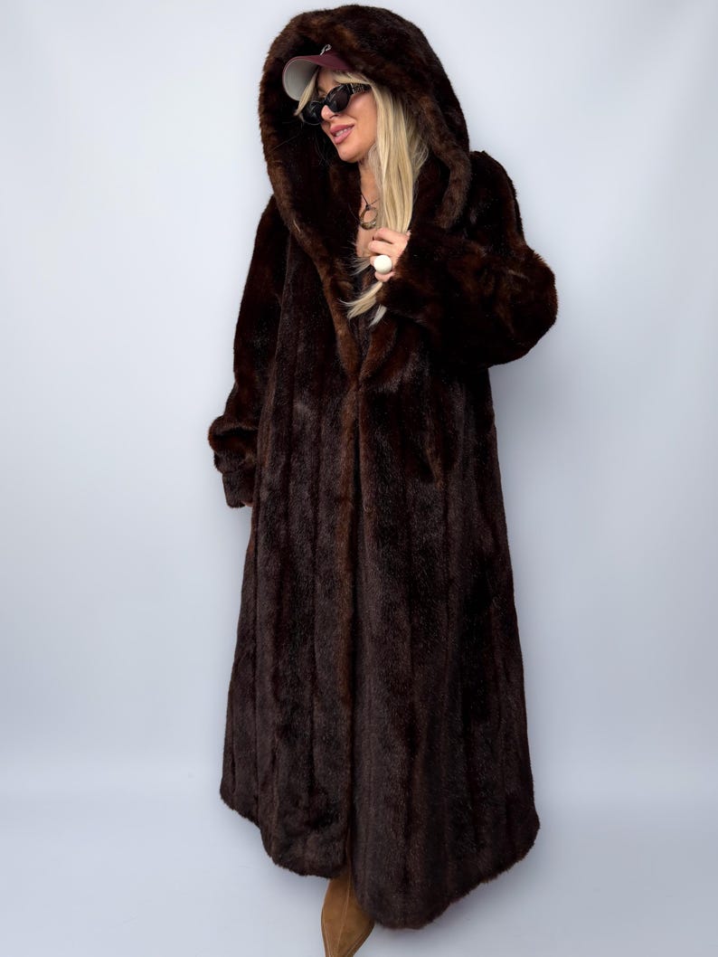 May include: A full-length, dark brown faux fur coat with a large hood. The coat has a long, flowing design and a plush texture. The hood is also lined with the same faux fur material. The coat is worn by a person wearing sunglasses and a baseball cap.