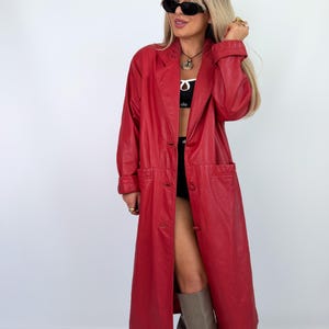 May include: A long, vibrant red leather coat with a classic lapel collar and button closure. The coat features long sleeves with cuff details and front pockets. The model is wearing black sunglasses and tall gray boots.