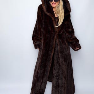 May include: A full-length, dark brown faux fur coat with a large hood. The coat is open in the front, revealing the lining. The coat has long sleeves and a soft, textured appearance. The model is wearing sunglasses and a cap.