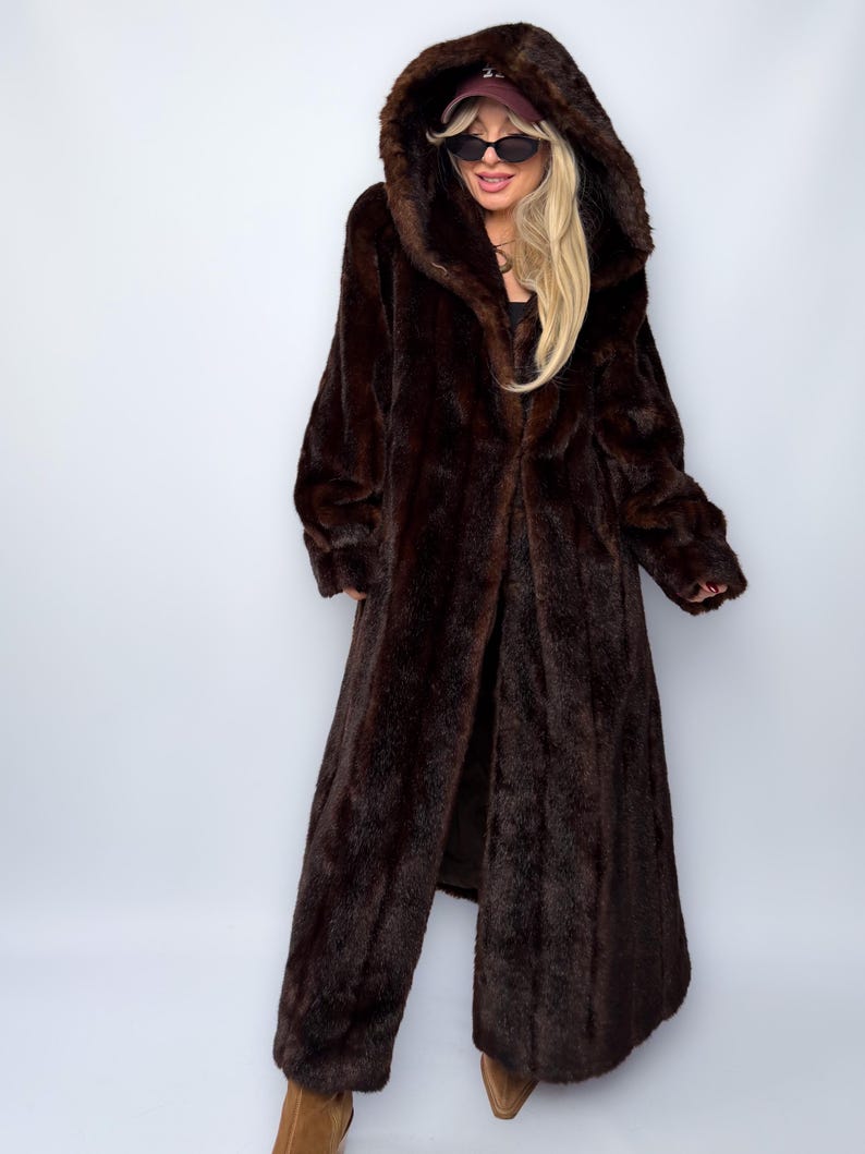 May include: A full-length, dark brown faux fur coat with a large hood. The coat is open in the front, revealing the lining. The coat has long sleeves and a soft, textured appearance. The model is wearing sunglasses and a cap.