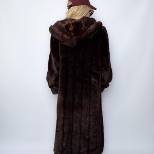 May include: A long, dark brown faux fur coat with a hood. The coat is floor-length and has a plush, textured appearance. The person is wearing a maroon baseball cap.