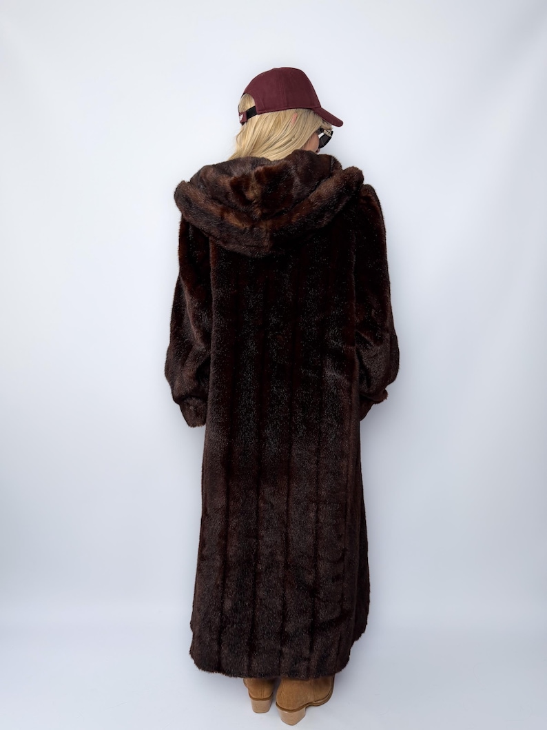 May include: A long, dark brown faux fur coat with a hood. The coat is floor-length and has a plush, textured appearance. The person is wearing a maroon baseball cap.