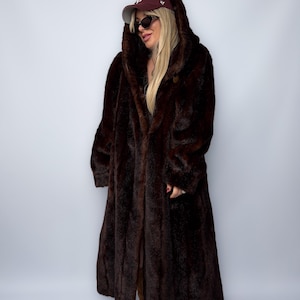 May include: A long, dark brown faux fur coat with a hood. The coat is floor-length and has a button closure. The person is wearing a maroon baseball cap and sunglasses. The coat appears to be warm and stylish.