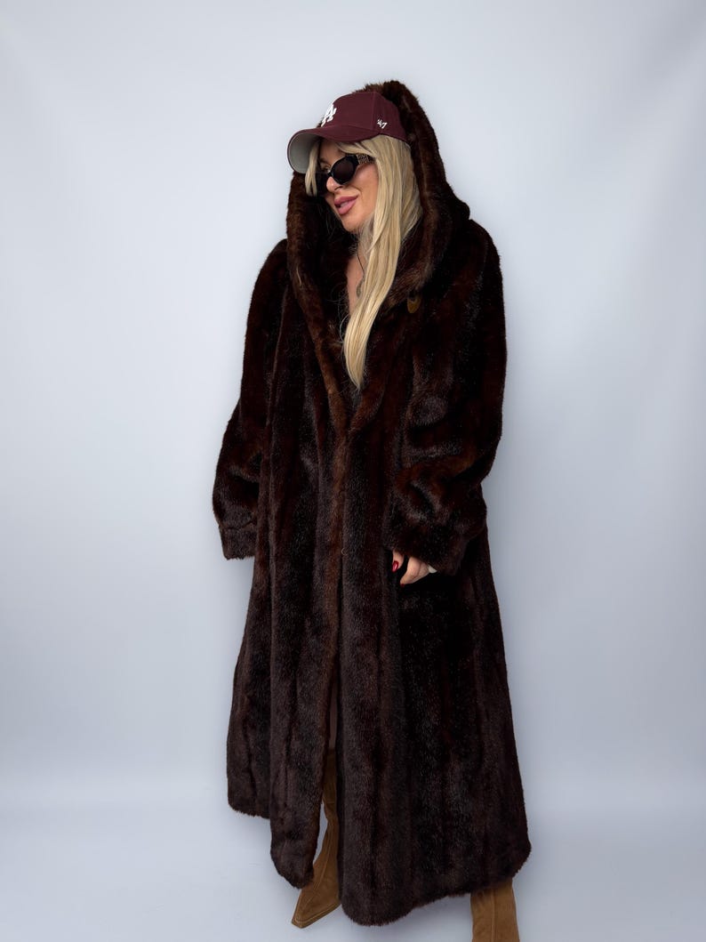 May include: A long, dark brown faux fur coat with a hood. The coat is floor-length and has a button closure. The person is wearing a maroon baseball cap and sunglasses. The coat appears to be warm and stylish.