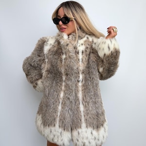 Può includere: Un cappotto in pelliccia sintetica a maniche lunghe con un motivo maculato marrone, grigio e bianco. Il cappotto presenta un colletto alto, una consistenza folta e ispida, e strisce verticali bianche sul davanti, con polsini e orlo bianchi.