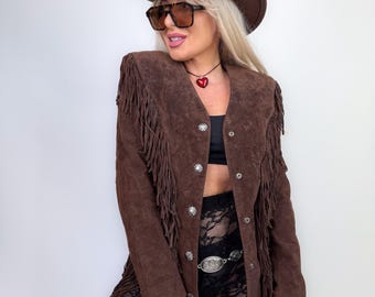 Puede incluir: Chaqueta de ante marrón con flecos, botones plateados y sombrero de vaquero a juego. La chaqueta tiene largos flecos en las mangas y el dobladillo. La persona lleva gafas de sol y un collar con forma de corazón.