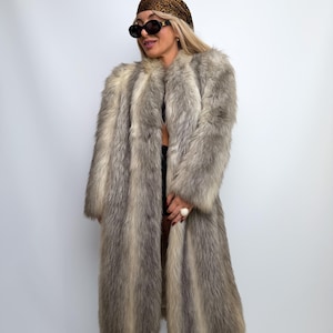 May include: A long, full-length faux fur coat featuring a variegated pattern of grey, white, and brown tones. The coat has long sleeves and a thick, textured pile. It is worn with a leopard print headscarf and black sunglasses.