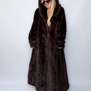 May include: A long, dark brown faux fur coat with a large hood. The coat is floor-length and has a classic design. The person is wearing a maroon baseball cap and sunglasses.