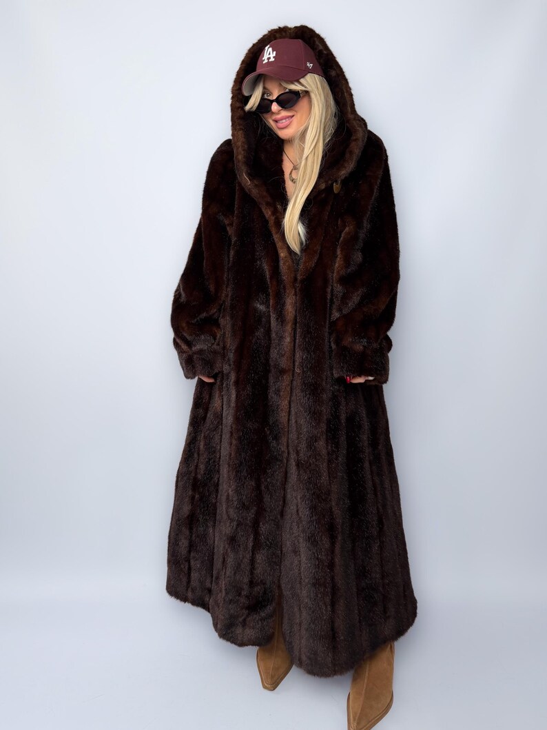 May include: A long, dark brown faux fur coat with a large hood. The coat is floor-length and has a classic design. The person is wearing a maroon baseball cap and sunglasses.