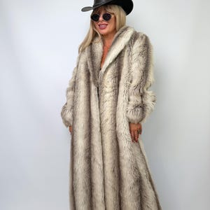 May include: A long, full-length faux fur coat featuring a vertical striped pattern in shades of cream, beige, and dark brown. The coat has long sleeves and a wide collar. A black cowboy hat and dark-tinted sunglasses are worn with the coat.