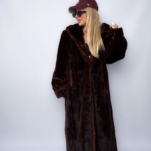 May include: A long, dark brown faux fur coat with a hooded design. The coat is floor-length and has a plush, textured appearance. The image also shows a maroon baseball cap and sunglasses.