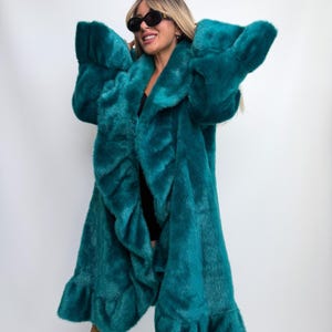 May include: A long, teal-coloured faux fur coat featuring a wide collar, long sleeves, and a ruffled trim detail along the front opening and the bottom hem. The coat has a thick, plush texture and is worn open over a simple black garment.