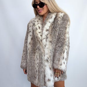 May include: A long-sleeved faux fur coat featuring a mottled pattern of brown, gray, and white tones. The coat has a wide lapel collar, a straight hemline, and a thick, textured pile throughout. The garment is worn with black sunglasses.