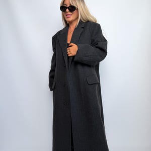 May include: A long, charcoal gray wool-blend overcoat featuring a notched lapel, structured shoulders, and a three-button front closure. The coat includes two side flap pockets and long sleeves, providing a full-length silhouette.