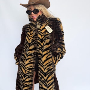 May include: A brown faux fur coat featuring a black tiger stripe pattern, a high collar, and a gold-toned button closure. The coat includes a white rectangular tag with the text RICHARD attached to the chest area, paired with a brown cowboy hat.
