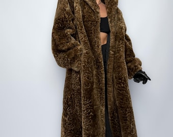 May include: A long, brown faux fur coat with a textured pattern. The coat has a wide collar and long sleeves. The coat is worn over a black top and trousers. The person is wearing black sunglasses and gloves.