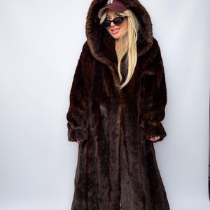 May include: A full-length, dark brown faux fur coat with a large hood. The coat has a long, flowing design and is worn over other clothing. The hood is up, and the coat appears to be made of a soft, plush material.