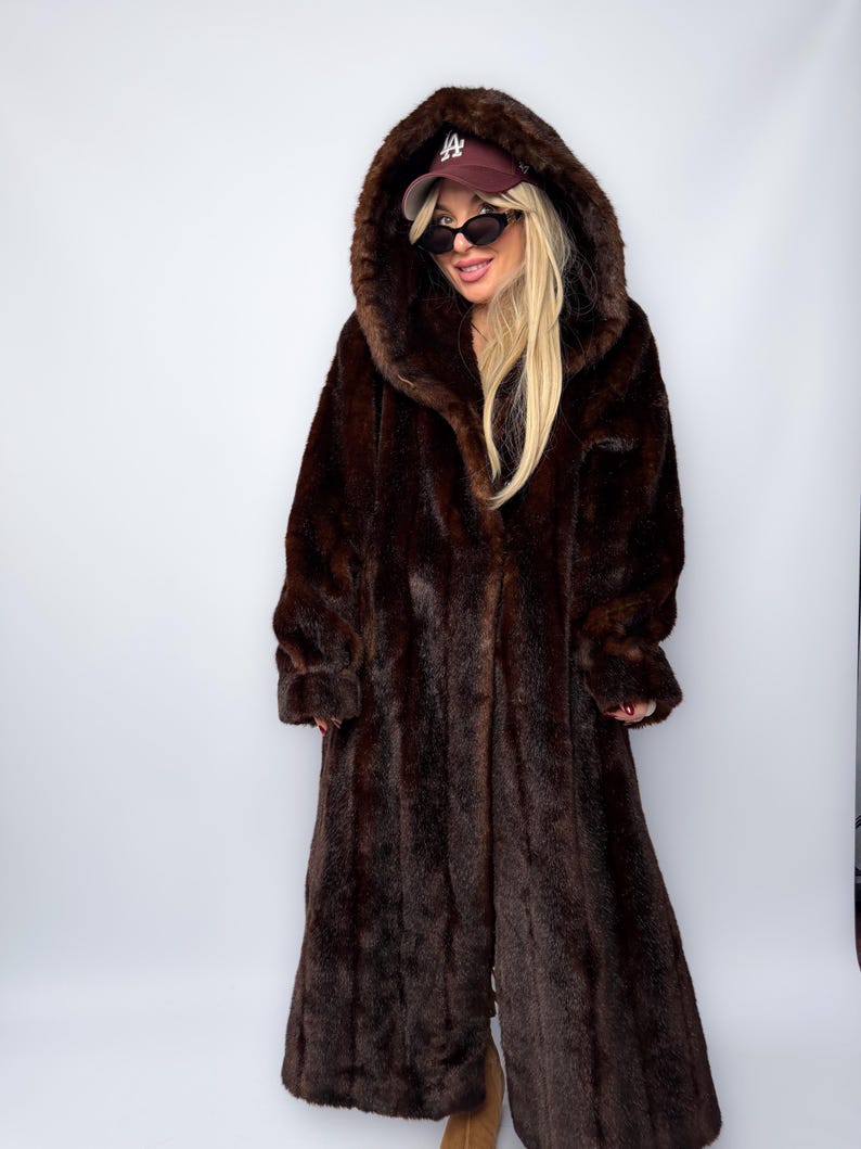 May include: A full-length, dark brown faux fur coat with a large hood. The coat has a long, flowing design and is worn over other clothing. The hood is up, and the coat appears to be made of a soft, plush material.