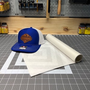 May include: A white and blue trucker hat with a brown leather patch that says "HYDBOND" on it. The hat is lying on a white sheet of paper with a pattern of diagonal lines. The paper is on a gray cutting mat.