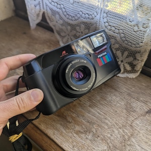 May include: Black Pentax Zoom Lens camera with a flash and a wrist strap. The camera has a zoom lens with the text "1:3.5-70mm TELE-MACRO". The camera has a multi-coloured stripe design on the side.