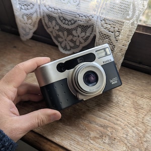 May include: A silver and black Konica Z-120 film camera with a zoom lens. The camera has a flash and the lens is marked 38mm - 120mm. The camera is resting on a wooden surface.