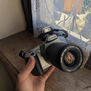 May include: A silver Canon EOS 3000 camera with a black lens and a built-in flash. The lens is a Canon Zoom Lens EF 28-90mm 1:4-5.6. The camera is held by a person.