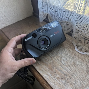 May include: Black Canon Prima Twin 35mm film camera with a built-in flash and lens. The camera has a black strap and is resting on a wooden surface. The camera has the Canon logo and Prima Twin printed on it.