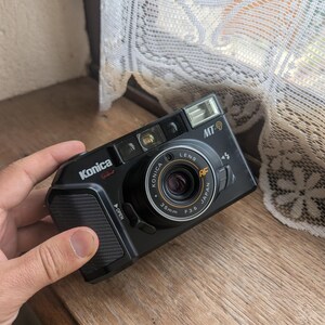 May include: Black Konica MT-9 35mm film camera with a built-in flash. The camera has a black body with the Konica logo and model name in white. The lens is labeled 35mm F3.5. The camera is held by a hand.