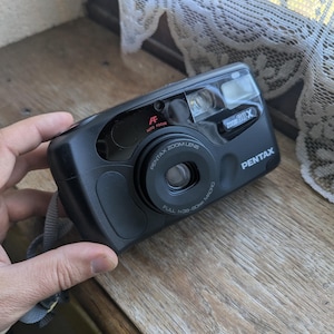 Może przedstawiać: Czarny aparat Pentax Zoom 90X z obiektywem zmiennoogniskowym i autofokusem. Aparat posiada wbudowaną lampę błyskową i pasek na nadgarstek. Obiektyw to Full 1:3.8-80mm Macro.