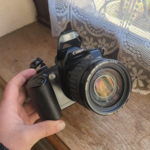 Puede incluir: Una cámara Canon EOS negra con un objetivo zoom, flash y el logotipo de Canon. La lente está etiquetada como 28-105mm 1:4-5.6. La cámara está sujeta por una mano.
