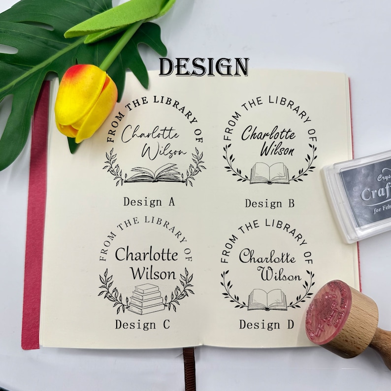 Peut inclure: Un tampon de biblioth&egrave;que personnalis&eacute; avec le texte "From the Library of Charlotte Wilson" dans un design circulaire, avec diff&eacute;rentes illustrations de livres. Le tampon est pr&eacute;sent&eacute; avec un manche en bois et un tampon encreur.