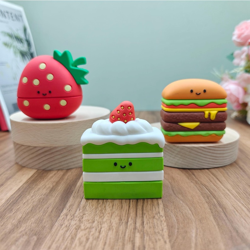 Puede incluir: Tres fundas de silicona con forma de alimentos y caras sonrientes. Una es una fresa roja con hojas verdes, otra es un pastel verde de varias capas con una fresa encima, y la tercera es una hamburguesa con queso, lechuga y queso amarillo.