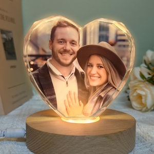 Op de afbeelding: Een hartvormige kristallen lampenvoet met een foto van een stel erin. De voet is van hout en heeft een warm, gloeiend licht.