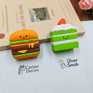 Puede incluir: Dos fundas de silicona para auriculares con forma de hamburguesa y pastel verde con una fresa encima. Debajo hay dos sellos de tinta negra: uno con un icono de unicornio y el texto Cartoo Davies, y otro con un dinosaurio y el nombre Oliver Smith.
