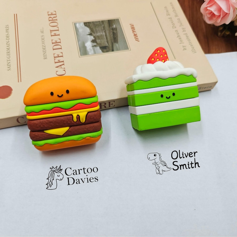Puede incluir: Dos fundas de silicona para auriculares con forma de hamburguesa y pastel verde con una fresa encima. Debajo hay dos sellos de tinta negra: uno con un icono de unicornio y el texto Cartoo Davies, y otro con un dinosaurio y el nombre Oliver Smith.