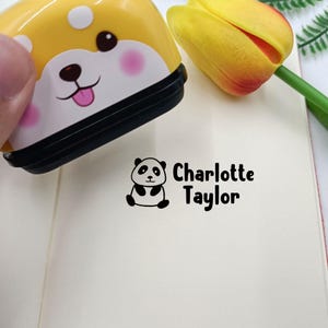 Puede incluir: Un sello autoentintable amarillo y blanco con un dise&ntilde;o de cara de perro. El sello crea una impresi&oacute;n de tinta negra sobre papel con una peque&ntilde;a ilustraci&oacute;n de un panda junto al texto impreso Charlotte Taylor en fuente sans-serif.
