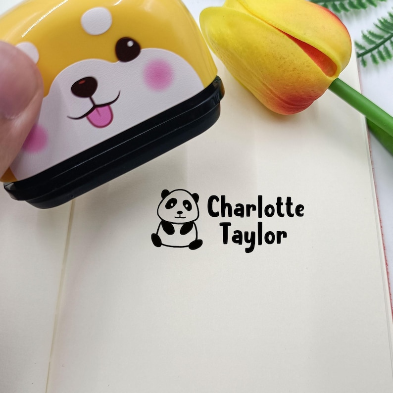 Puede incluir: Un sello autoentintable amarillo y blanco con un dise&ntilde;o de cara de perro. El sello crea una impresi&oacute;n de tinta negra sobre papel con una peque&ntilde;a ilustraci&oacute;n de un panda junto al texto impreso Charlotte Taylor en fuente sans-serif.