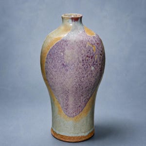 May include: A ceramic vase with a narrow neck and a rounded body, featuring a mottled purple glaze on the front, surrounded by areas of tan, beige, and light gray. The base of the vase is unglazed, showing a textured, reddish-brown clay surface.