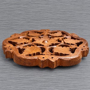 May include: A circular wooden trivet featuring an intricate, hand-carved openwork design of multiple maple leaves arranged in a symmetrical pattern. The natural brown wood grain is visible throughout the piece, which has a flat base and edges.