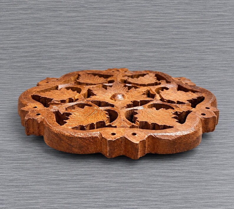 May include: A circular wooden trivet featuring an intricate, hand-carved openwork design of multiple maple leaves arranged in a symmetrical pattern. The natural brown wood grain is visible throughout the piece, which has a flat base and edges.