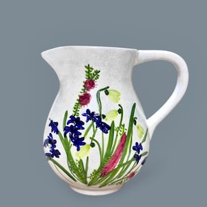 May include: A white ceramic pitcher featuring a hand-painted botanical design. The artwork displays vibrant blue hyacinth flowers, pale yellow snowdrops, and tall green stems with textured pink and red accents against a smooth, off-white surface.