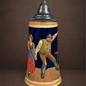 May include: A ceramic beer stein with a dark blue background featuring a raised relief of a person in a green vest and brown trousers dancing next to another person in a red skirt. The stein has a pointed, embossed metal lid and a tan base.
