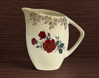 May include: A cream-colored ceramic pitcher featuring a central floral motif of deep red roses with green leaves. The upper rim is decorated with a delicate gold-toned floral pattern. The pitcher has a single handle and a curved pouring spout.