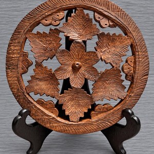 May include: A circular brown wooden trivet featuring an intricate openwork carving of a central six-petaled flower surrounded by six detailed leaves. The outer rim has a textured, grooved pattern, and the piece is displayed on a black stand.