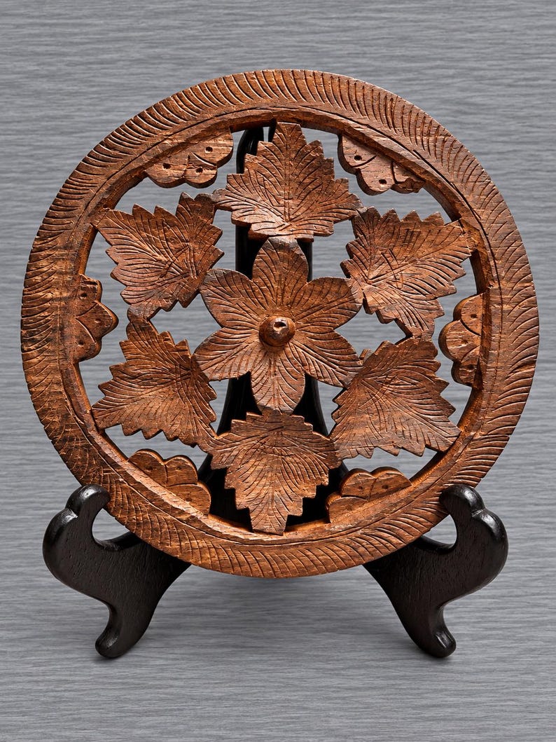 May include: A circular brown wooden trivet featuring an intricate openwork carving of a central six-petaled flower surrounded by six detailed leaves. The outer rim has a textured, grooved pattern, and the piece is displayed on a black stand.