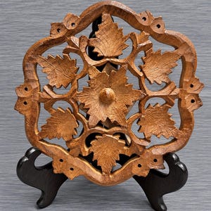 May include: A circular brown wooden trivet featuring an intricate openwork carving of a central flower surrounded by six detailed leaves. The piece has a scalloped outer border with small circular cutouts and rests on a black display stand.
