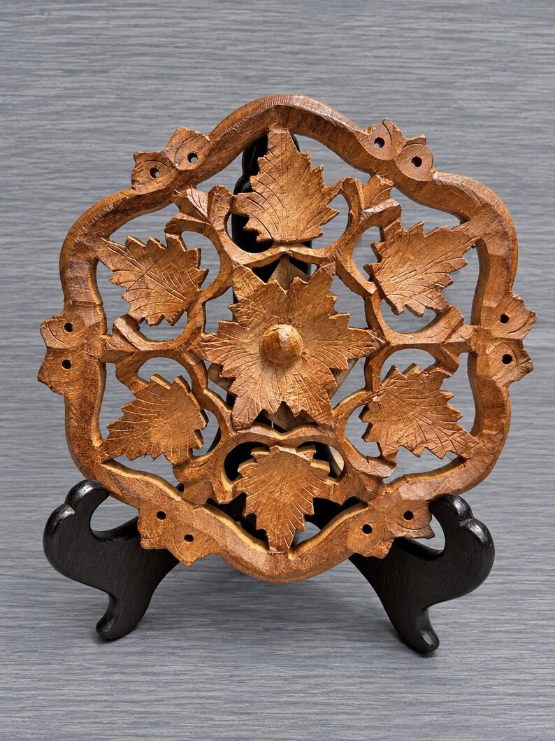 May include: A circular brown wooden trivet featuring an intricate openwork carving of a central flower surrounded by six detailed leaves. The piece has a scalloped outer border with small circular cutouts and rests on a black display stand.
