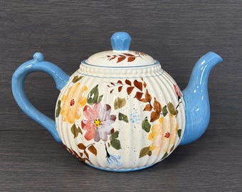 May include: A ceramic teapot with a cream-colored, ribbed body featuring painted yellow and pink flowers, green leaves, and brown branches. The handle, spout, and lid knob are light blue. The surface has a speckled finish throughout.